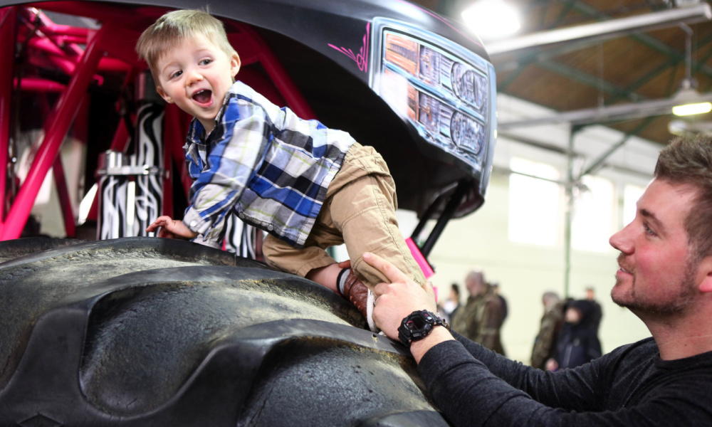 A small child around 3 years old is boosted onto the massive tire of a monster truck at the 2018 Kids Expo.