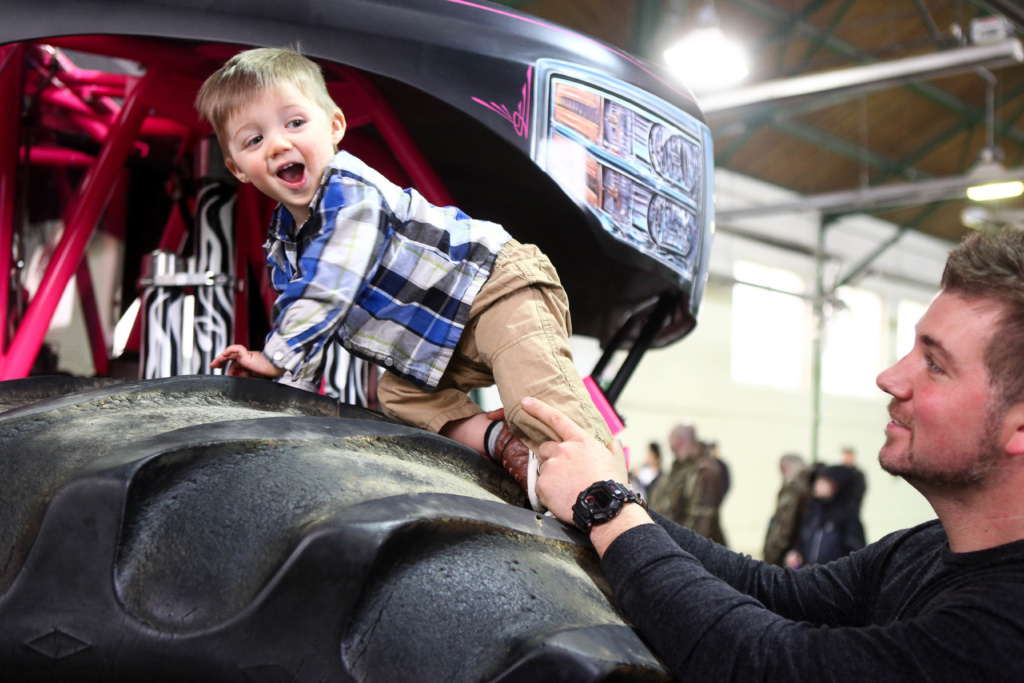 A small child around 3 years old is boosted onto the massive tire of a monster truck at the 2018 Kids Expo.