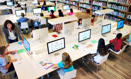 A stock photo shows a top-down angle of a library, where abut 10 young students sit around a table, each in front of their own computer.