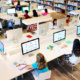 A stock photo shows a top-down angle of a library, where abut 10 young students sit around a table, each in front of their own computer.