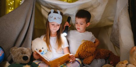 Two children inside fort reading a book with a flashlight
