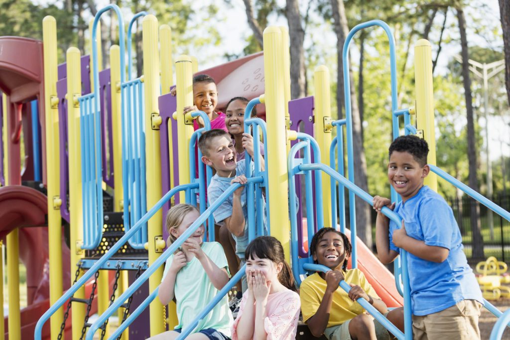 A multi-ethnic group of seven children, 8 to 10 years old, playing together on a playground. They are all hanging out on the play equipment and are smiling and laughing. The girl with light brown hair, on the left side, has down syndrome.