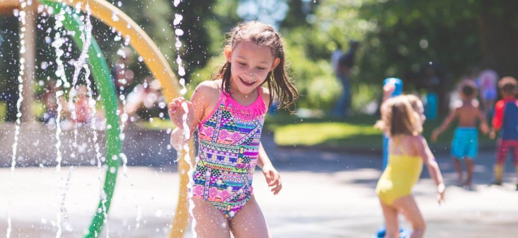 A young girl in a swimsuit is running underneath one of the sprinklers while other kids play at the splash pad.