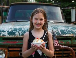 Young girl poses in front of old car with bowl of ice cream.