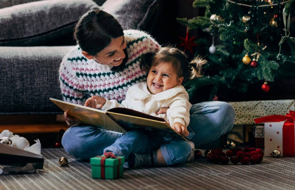 Smiling mother reading a Christmas story to her toddler daughter at home.